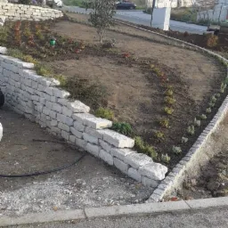 Wznoszenie kamiennego murku oporowego na zboczu, z widocznymi warstwami ułożonych bloków i świeżo posadzoną roślinnością, w tle fragment zabudowy mieszkalnej i drogi.