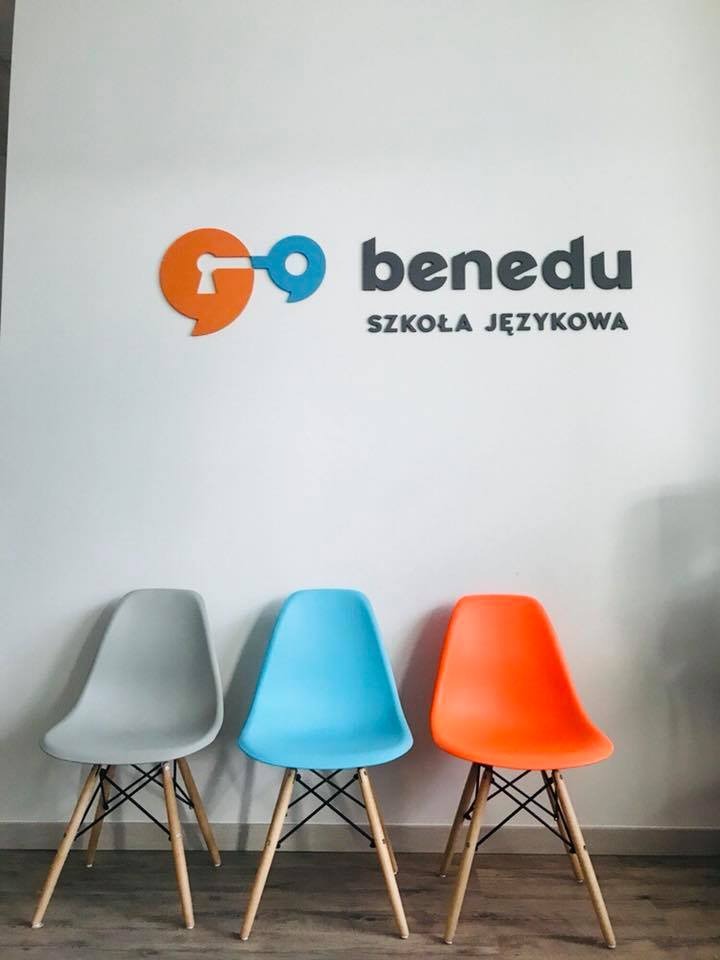 Logo 'benedu Szkoła Językowa' na białej ścianie, poniżej trzy krzesła w kolorach: szarym, błękitnym i pomarańczowym.