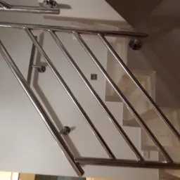 Polerowana, metalowa konstrukcja zabezpieczająca schody, z widocznymi mocowaniami do ściany, w neutralnej kolorystyce wnętrza.