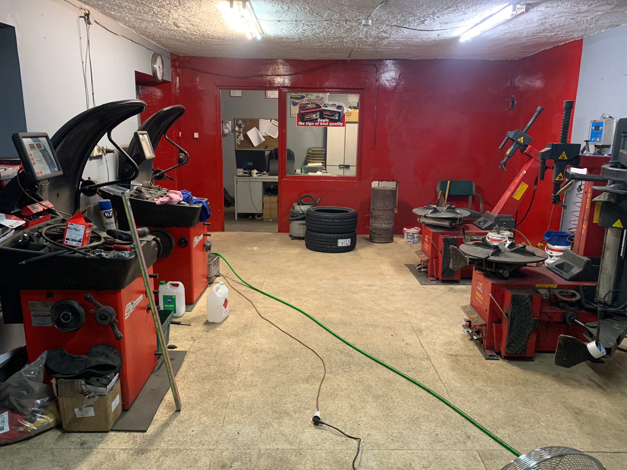 Wnętrze warsztatu wulkanizacyjnego z dwoma wyważarkami do kół, dwiema montażownicami opon oraz stosem opon w tle, ściany pomalowane na czerwono.