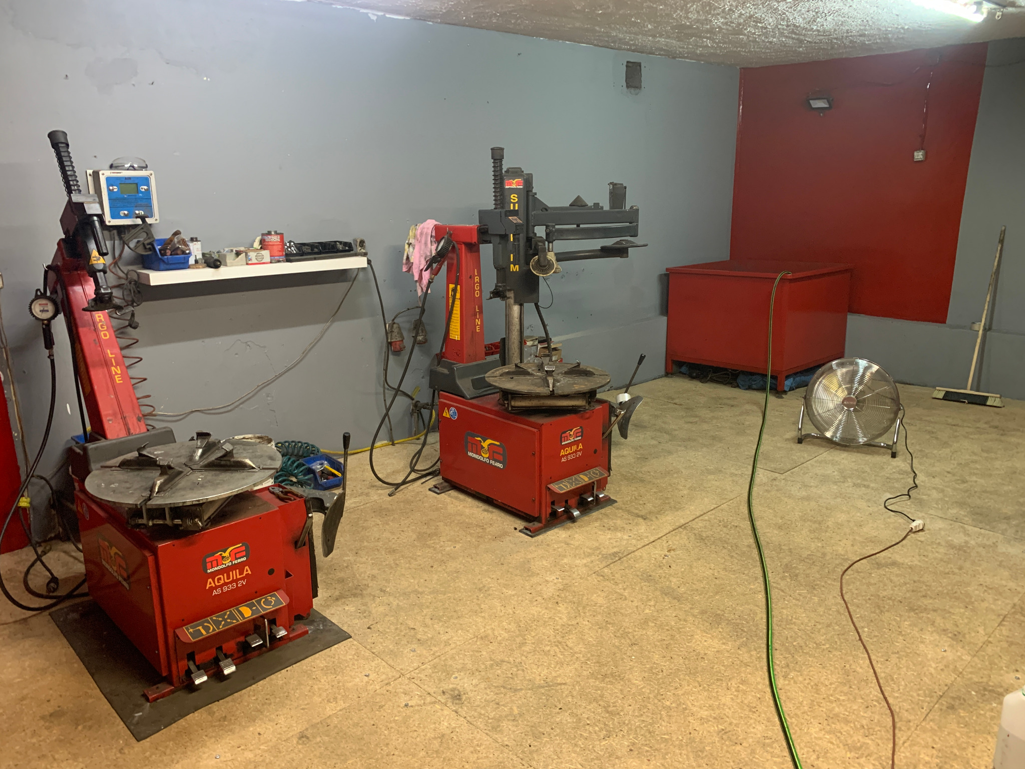 Wnętrze warsztatu z dwoma czerwonymi urządzeniami do montażu opon, widoczny wentylator i narzędzia na półce, ściany szare i czerwone.