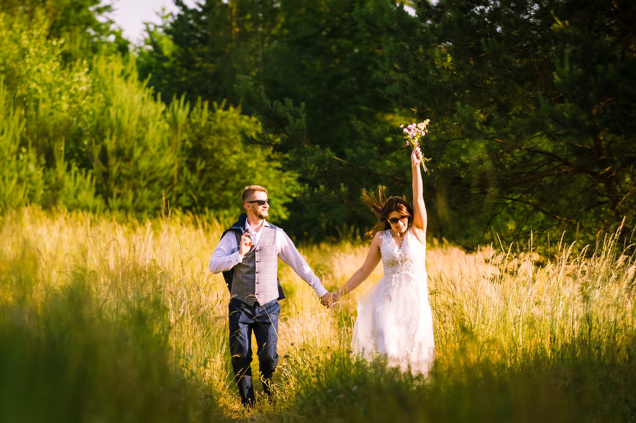Para młoda w okularach przeciwsłonecznych biegnie trzymając się za ręce przez pole z wysoką trawą; panna młoda unosi bukiet kwiatów w górę.