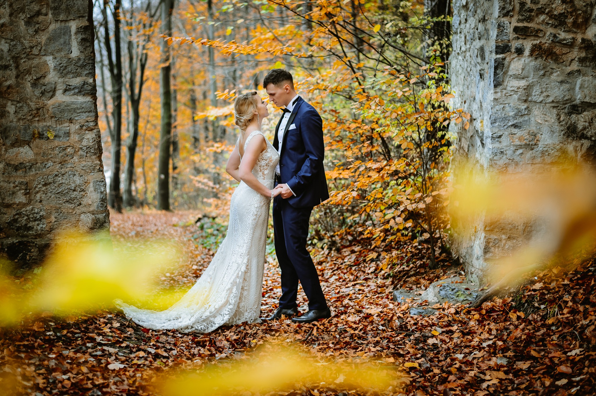 Para młoda trzymająca się za ręce w jesiennym lesie, panna młoda w koronkowej sukni, pan młody w granatowym garniturze, tło stanowią kamienne ruiny i opadłe liście.