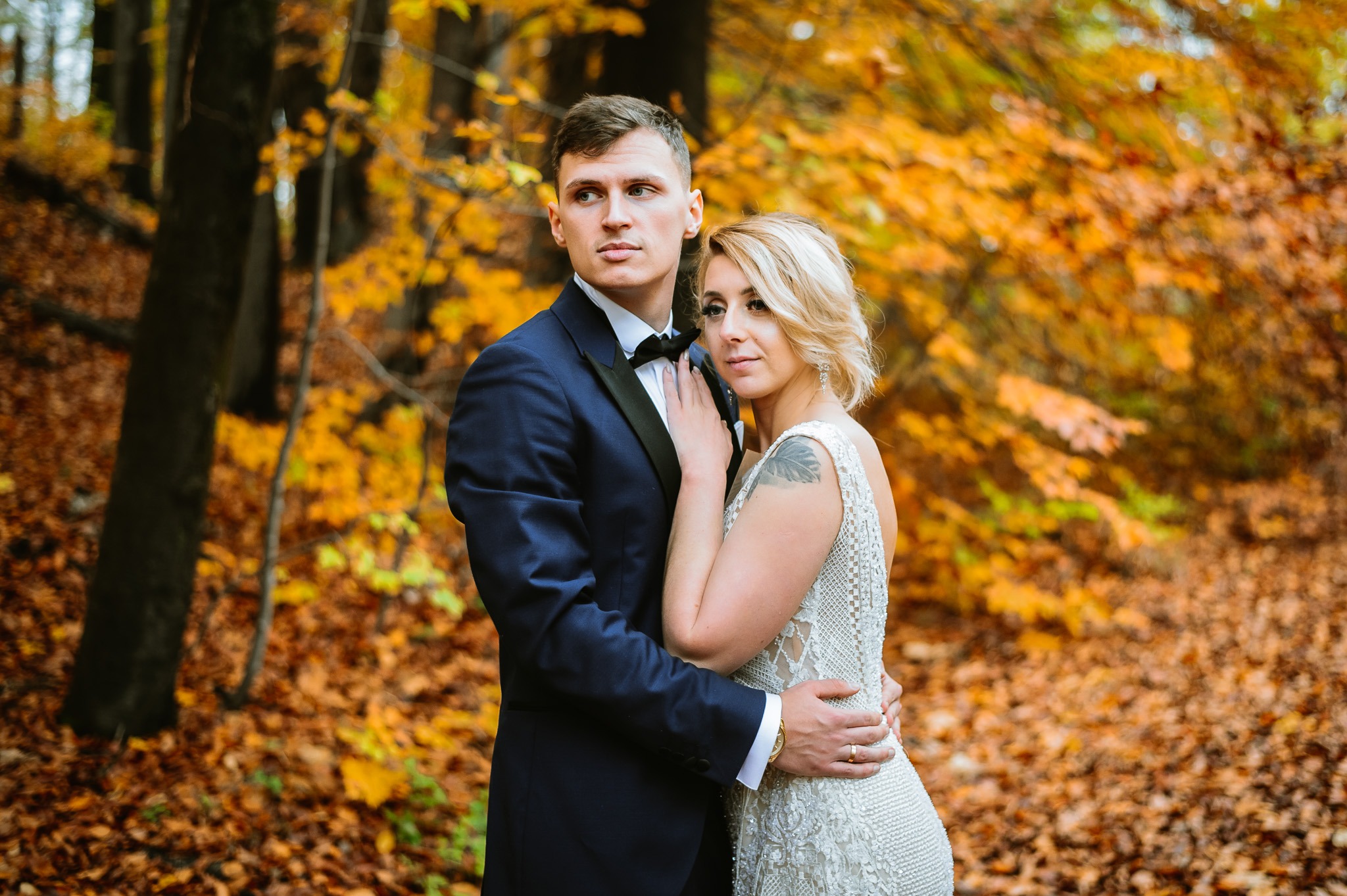 Para młoda w jesiennym lesie: mężczyzna w granatowym smokingu z muchą obejmuje kobietę w białej, zdobionej sukni z tatuażem na ramieniu, na tle drzew z pomarańczowymi liśćmi.