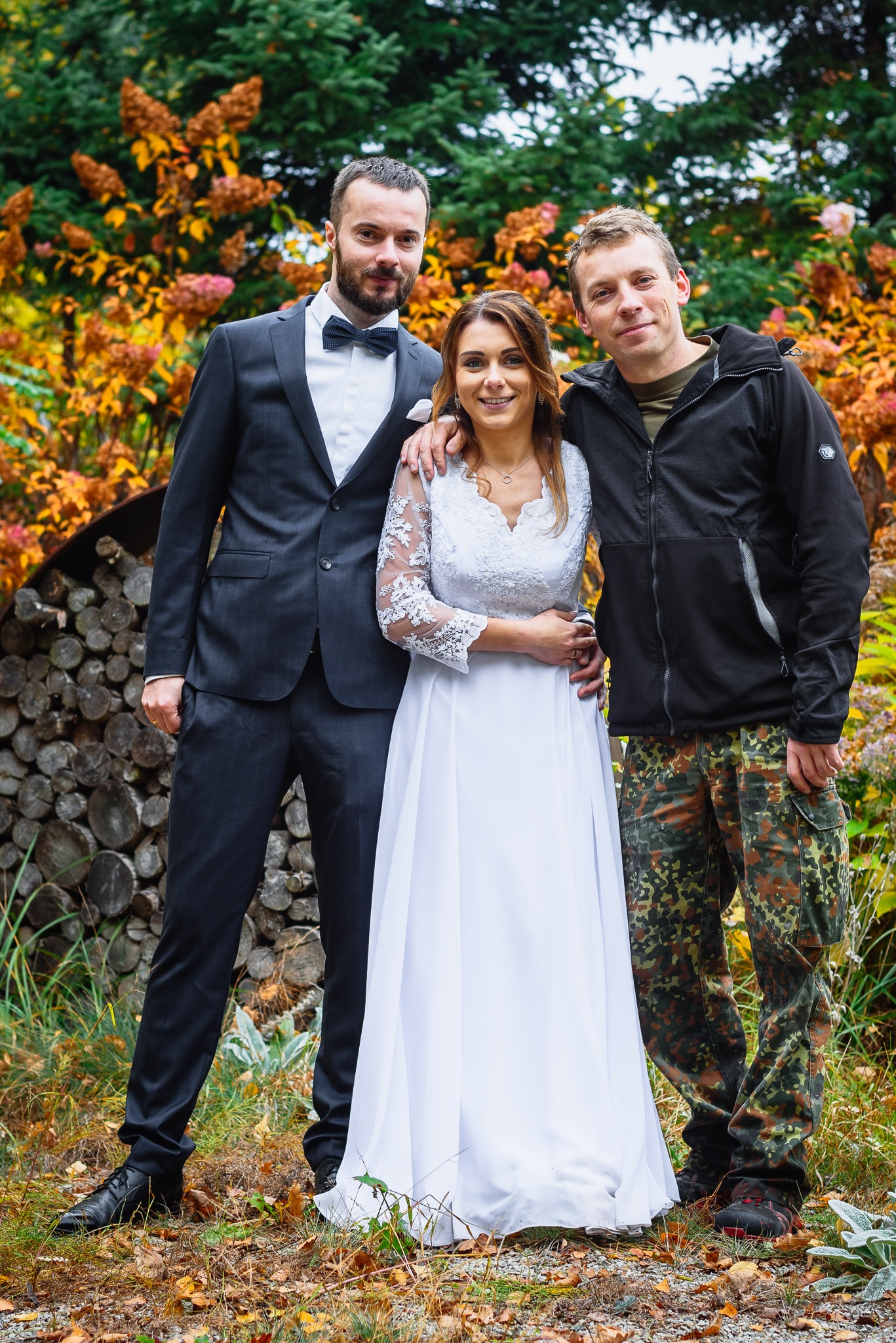 Panna młoda w białej sukni ślubnej z koronkowymi rękawami pozuje z dwoma mężczyznami na tle jesiennego ogrodu i stosu drewna.