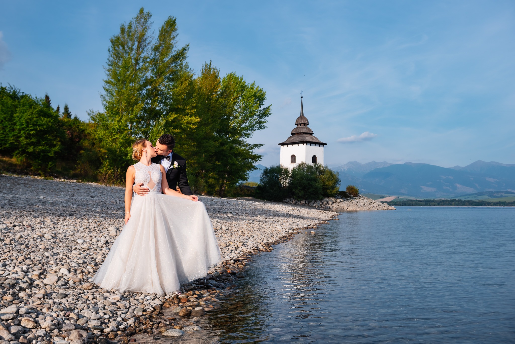 Pocałunek pary młodej na kamienistej plaży jeziora z widokiem na białą wieżę kościoła i odległe góry.