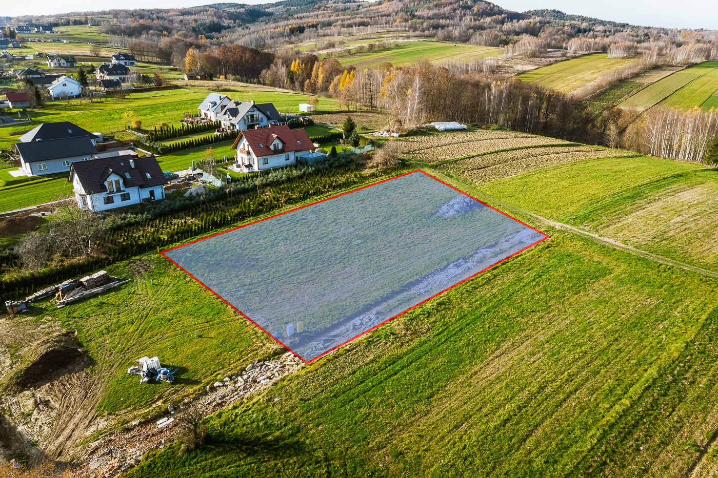 Działka budowlana z zaznaczonym obrysem, widok z drona na zielone pola, zabudowania mieszkalne w tle, jesienne drzewa w oddali.