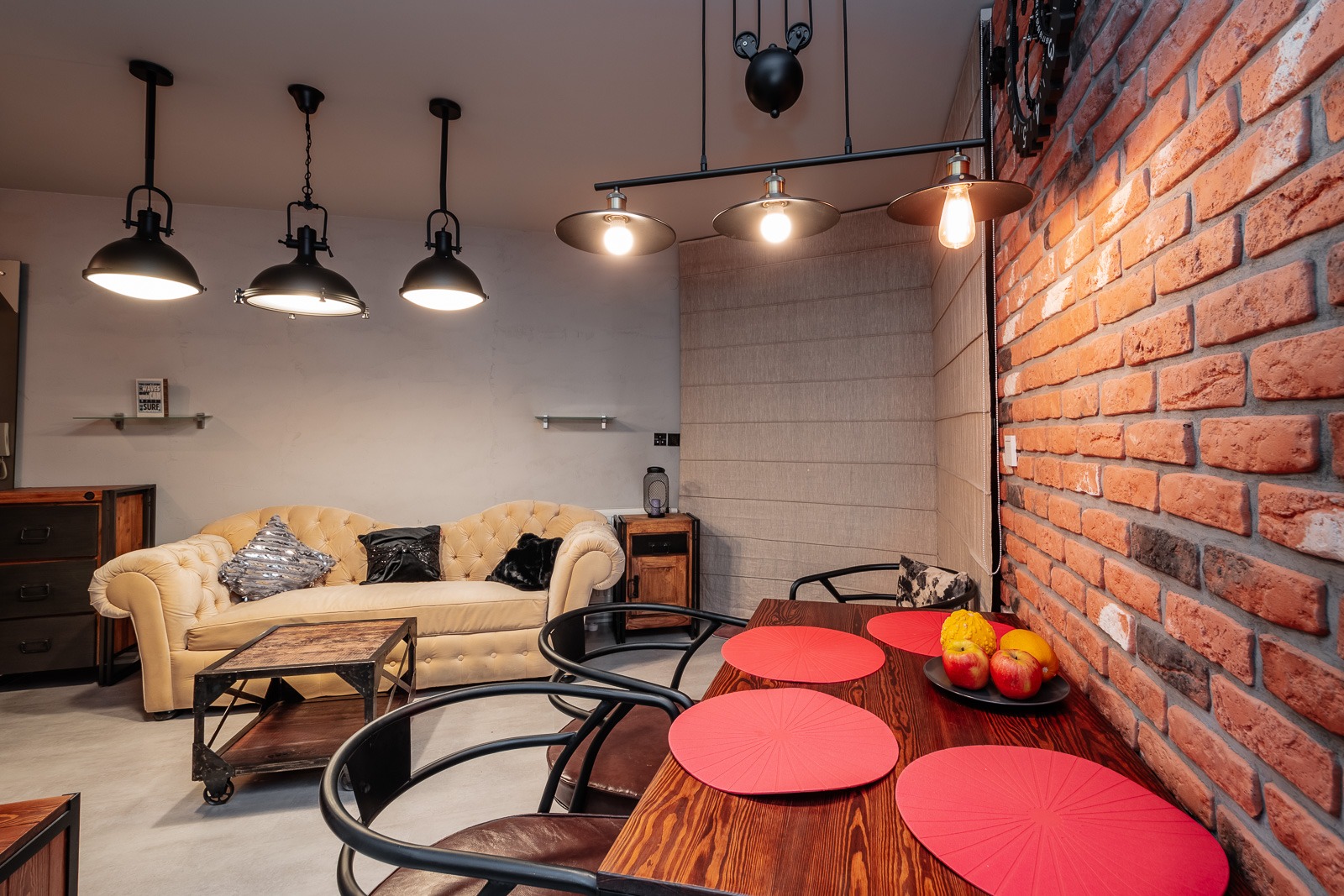 Loftowe wnętrze z ceglaną ścianą, drewnianym stołem z czerwonymi podkładkami, beżową sofą i industrialnym oświetleniem.