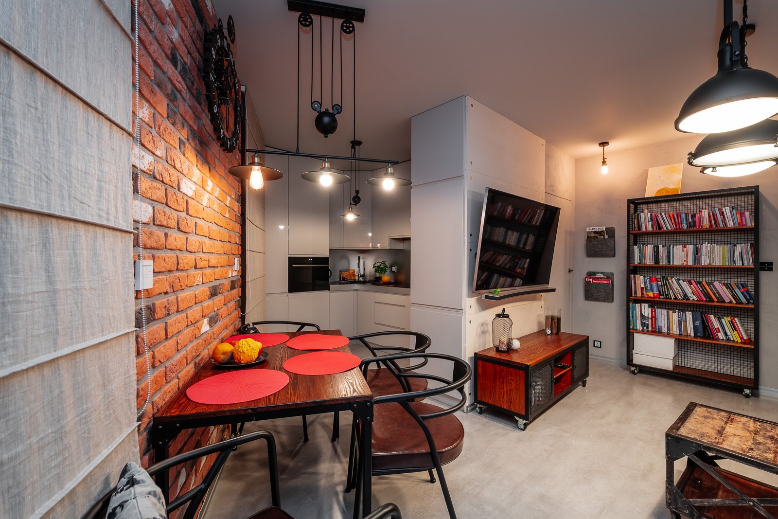 Nowoczesne wnętrze mieszkania z ceglaną ścianą, drewnianym stołem z czerwonymi podkładkami, otwartą kuchnią w tle i telewizorem z półką zawieszonym na ścianie.