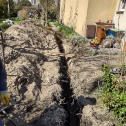 Wykopany wąski rów w ziemi wzdłuż ściany budynku, z widoczną rurą w oddali, podpartą drewnianą deską; na pierwszym planie narzędzie z żółtą rękojeścią i pracownik w rękawicach.