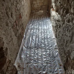 Instalacja ogrzewania podłogowego w trakcie montażu w Łazach, widok z góry na rozłożone rury na folii izolacyjnej, surowe kamienne ściany w tle.