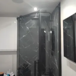 Kabina prysznicowa z czarnymi marmurkowymi ścianami i czarną ramą, widoczna w lustrze umywalka z przyborami toaletowymi.