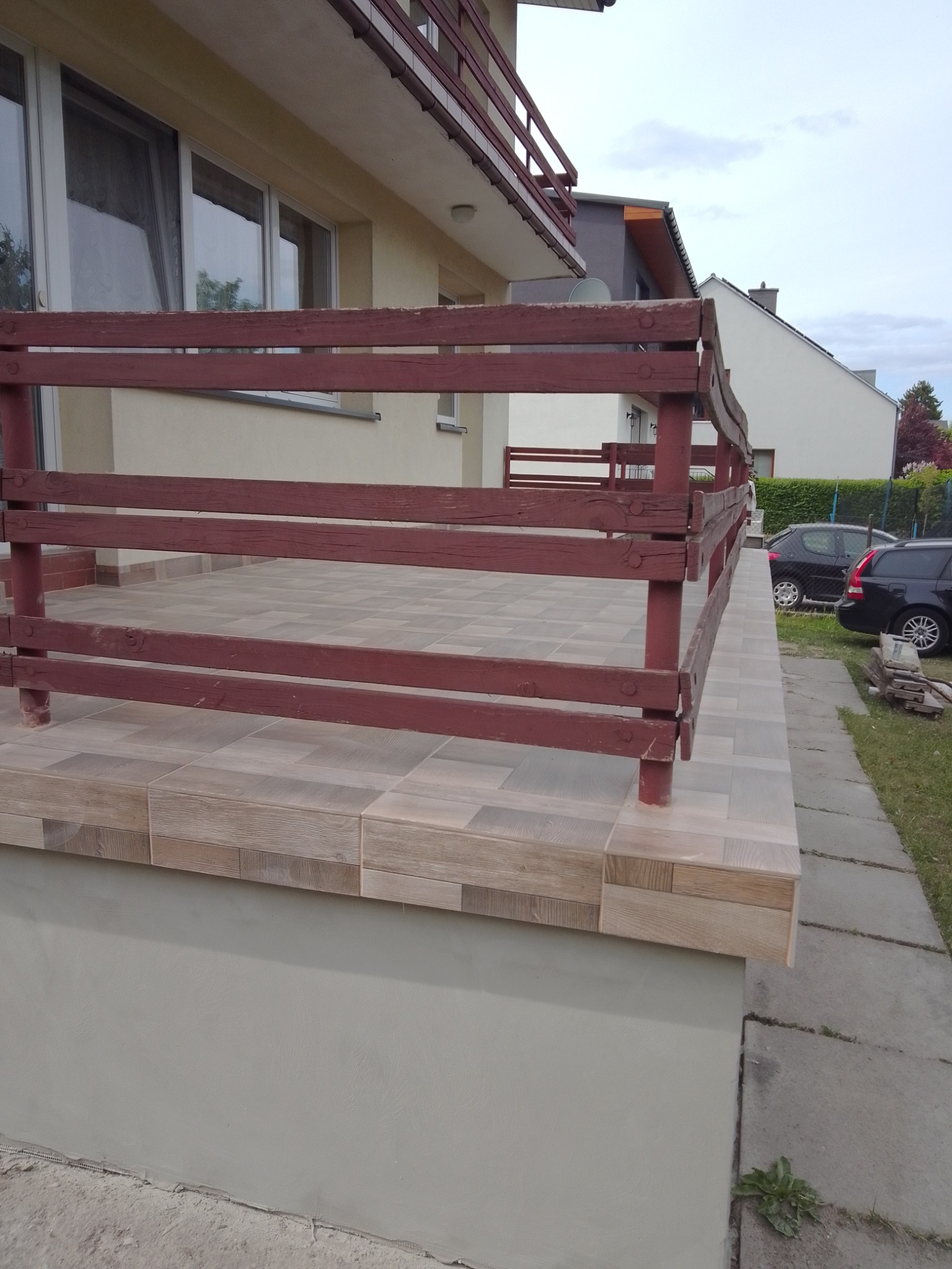Wykończony taras z brązową drewnianą barierką i płytkami imitującymi drewno, widok na dom i zaparkowane samochody.