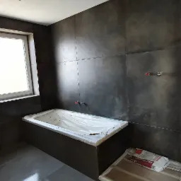 Łazienka w trakcie remontu: położone ciemne płytki na ścianach i podłodze, wanna zabezpieczona folią, widoczne puszki elektryczne i opakowanie kleju do płytek.