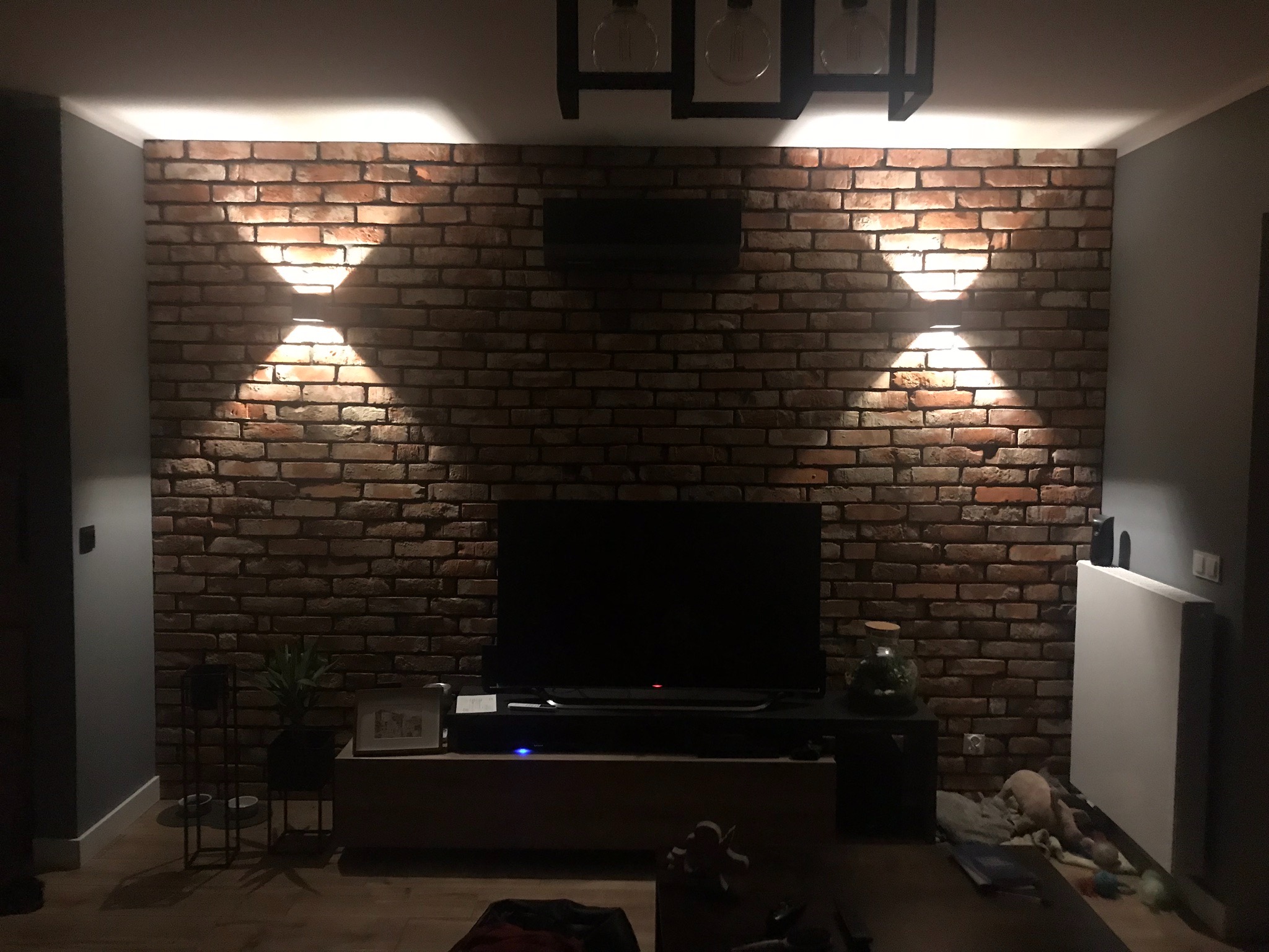 Ściana z cegły klinkierowej w salonie, oświetlona dwoma kinkietami tworzącymi efektowne stożki światła, nad telewizorem dekoracyjna czarna lampa sufitowa, widoczne elementy wystroju wnętrza.