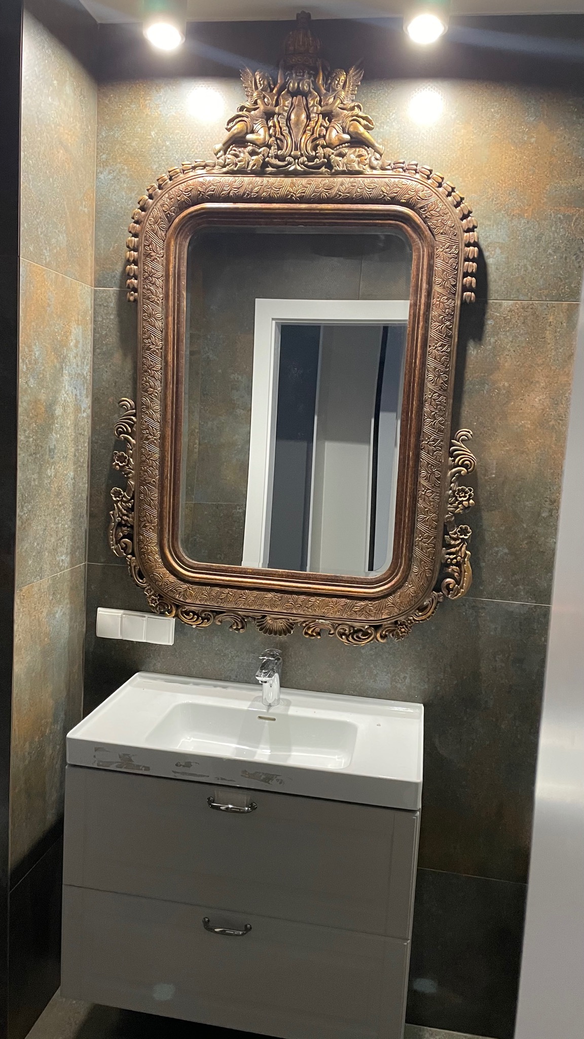 Umywalka z szafką w łazience, nad nią ozdobne, złote lustro z motywem aniołów, ściany wyłożone płytkami w odcieniach szarości i brązu.