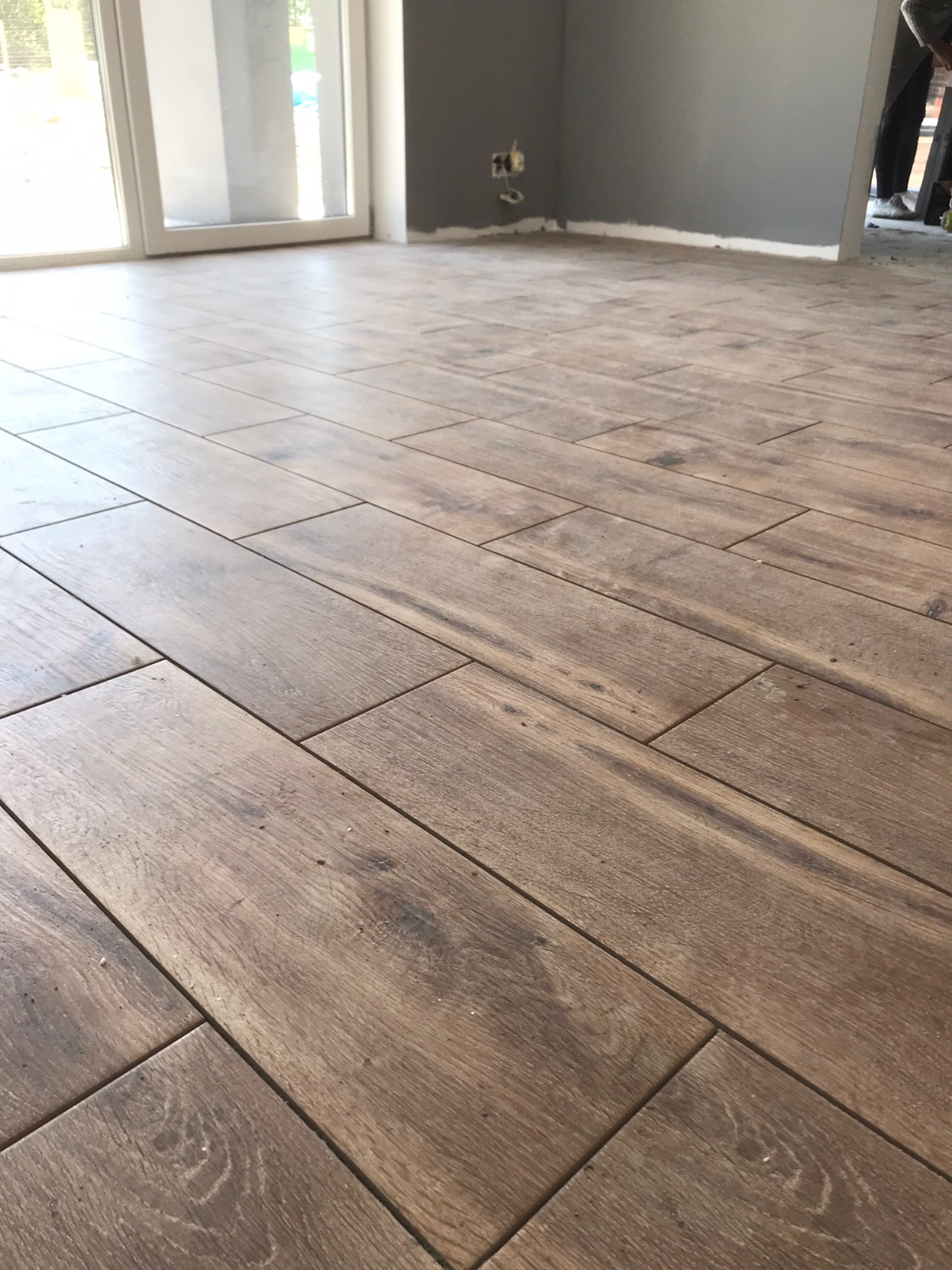 Ułożona podłoga z płytek imitujących drewno, widoczna szara ściana i białe listwy przypodłogowe, w tle okno balkonowe.