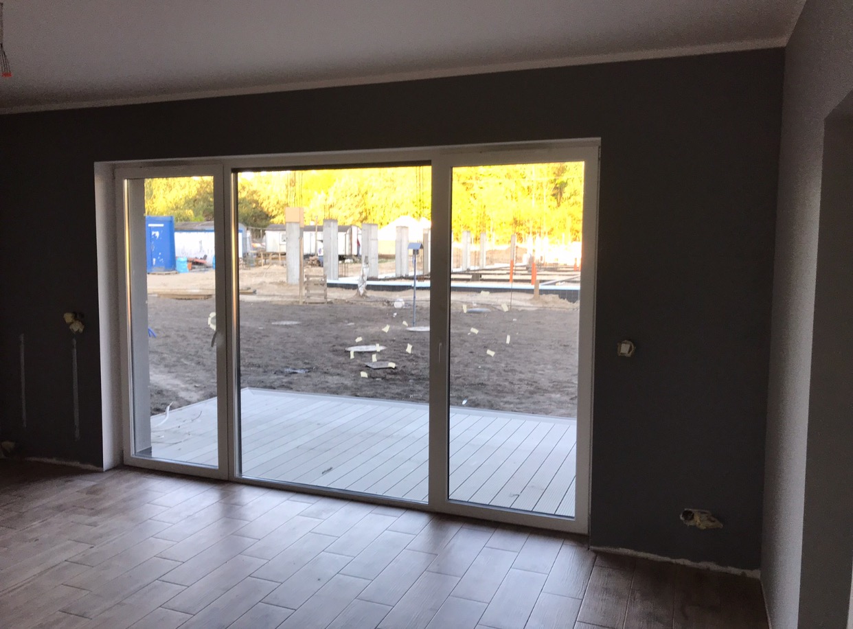 Wnętrze pokoju z dużym przesuwnym oknem balkonowym, widok na plac budowy z elementami konstrukcji i zapleczem budowlanym, brązowa podłoga imitująca drewno, szare ściany z wystającymi przewodami...