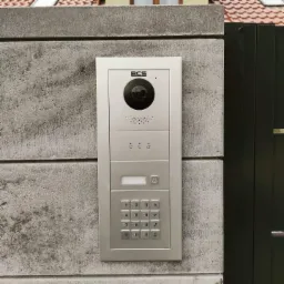 Srebrny panel domofonowy BCS z kamerą i klawiaturą numeryczną, zamontowany na betonowym słupku ogrodzeniowym obok czarnej furtki.