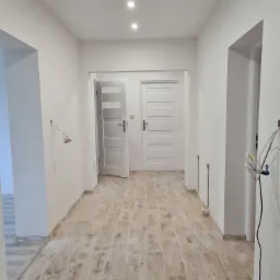 Korytarz w trakcie remontu: świeżo pomalowane białe ściany, ułożona podłoga imitująca drewno, widoczne niedokończone instalacje elektryczne i hydrauliczne, białe drzwi wewnętrzne z poziomymi panelami.