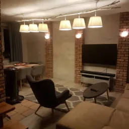 Salon z ceglanymi filarami, oświetleniem na gałęzi i nowoczesnym wystrojem, w tym szary fotel i beżowa sofa.