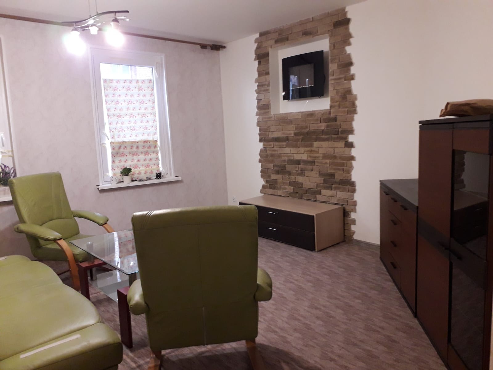 Salon z dekoracyjną ścianą z cegły wokół kominka elektrycznego, zielonymi fotelami i kanapą, oraz brązową podłogą imitującą drewno.