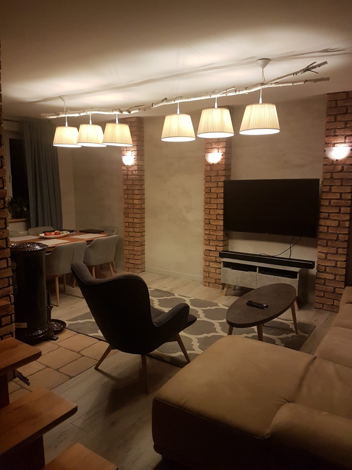 Salon z ceglanymi filarami, oświetleniem na gałęzi i nowoczesnym wystrojem, w tym szary fotel i beżowa sofa.