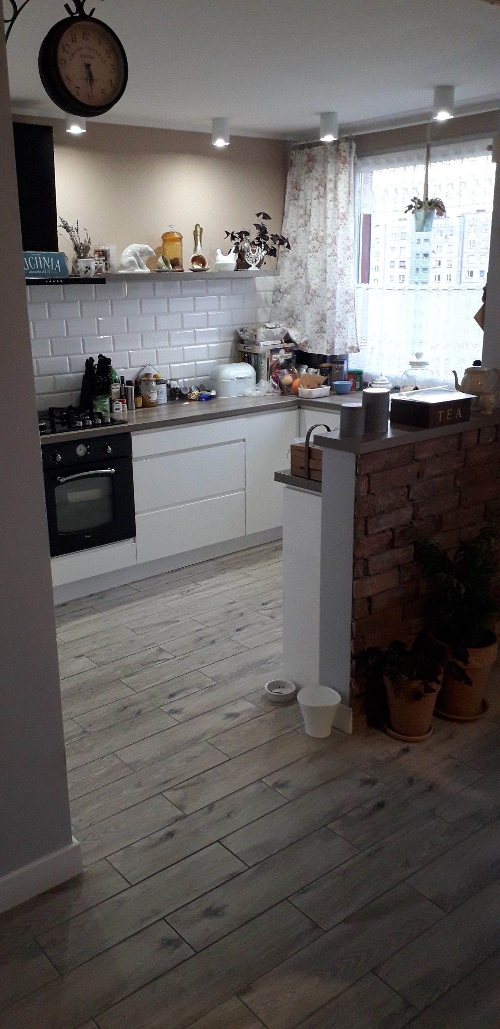 Jasna kuchnia z białymi meblami, szarą podłogą imitującą drewno i ceglanym elementem dekoracyjnym, widok na okno z kwiatami.