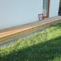 Parkiety Schody Tarasy Mirosław Rokicki - Jasny taras z desek przy białej ścianie domu, z czerwoną ramką okienną opartą o ścianę. Widok na trawnik w słoneczny dzień.