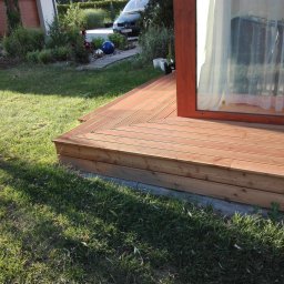 Parkiety Schody Tarasy Mirosław Rokicki - Jasny taras drewniany z desek ułożonych w jodełkę, przylegający do budynku z oknem. W tle trawnik, roślinność i zaparkowany samochód.