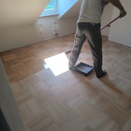 Parkiety Schody Tarasy Mirosław Rokicki - Parkiet w jodełkę po cyklinowaniu, częściowo polakierowany. Widoczny pracownik z wałkiem i kuwetą. Poddasze z oknem dachowym. Jasne, naturalne światło.