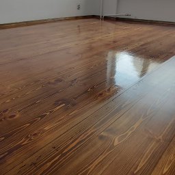 Parkiety Schody Tarasy Mirosław Rokicki - Błyszcząca, świeżo polakierowana podłoga z desek w odcieniach brązu, odbijająca światło z okna. Widoczne detale usłojenia drewna i białe ściany w tle.