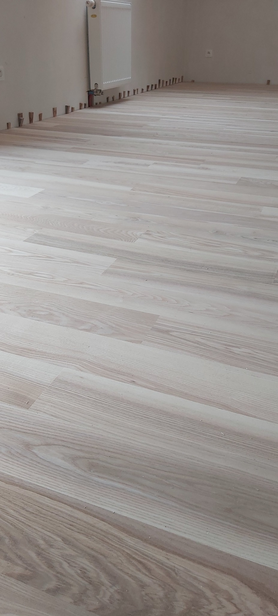 Jasne panele podłogowe ułożone w pokoju, widoczne kołki dystansowe przy ścianie i grzejnik. Delikatny wzór drewna, świeżo po montażu, gotowe do wykończenia.