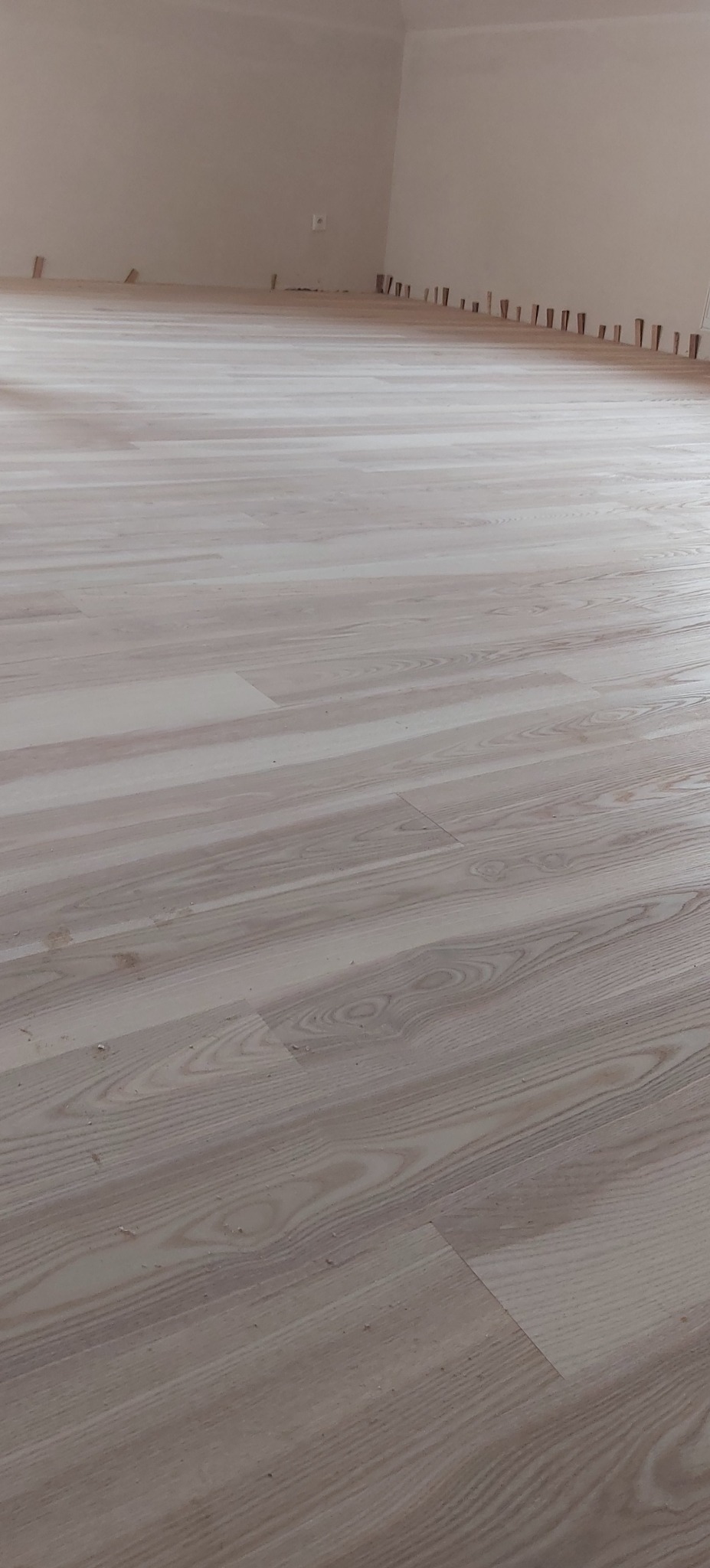 Jasne panele podłogowe z widocznym wzorem drewna, świeżo ułożone w pustym pomieszczeniu. Listwy przypodłogowe jeszcze nie zamontowane, widoczne kołki dystansowe.
