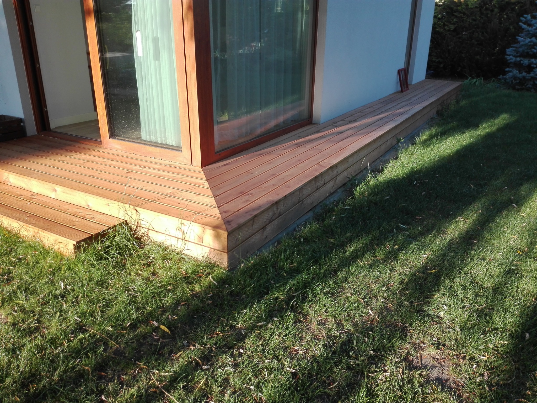 Drewniany taras przylegający do budynku z dużym oknem, otoczony trawnikiem. Widoczne detale konstrukcyjne i naturalne oświetlenie.