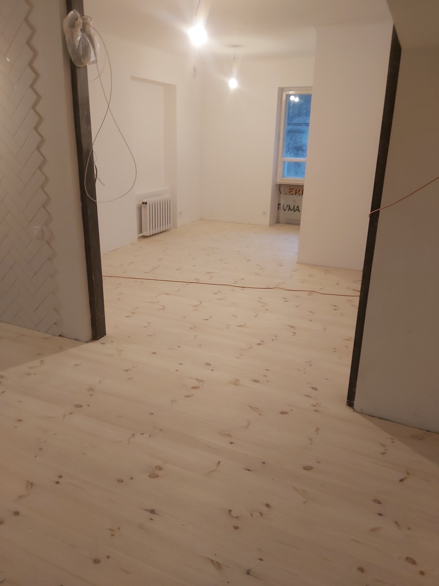 Jasne, świeżo położone deski podłogowe z widocznymi sękami, w pustym pokoju z białymi ścianami i grzejnikiem. Widok z perspektywy wejścia do pomieszczenia.