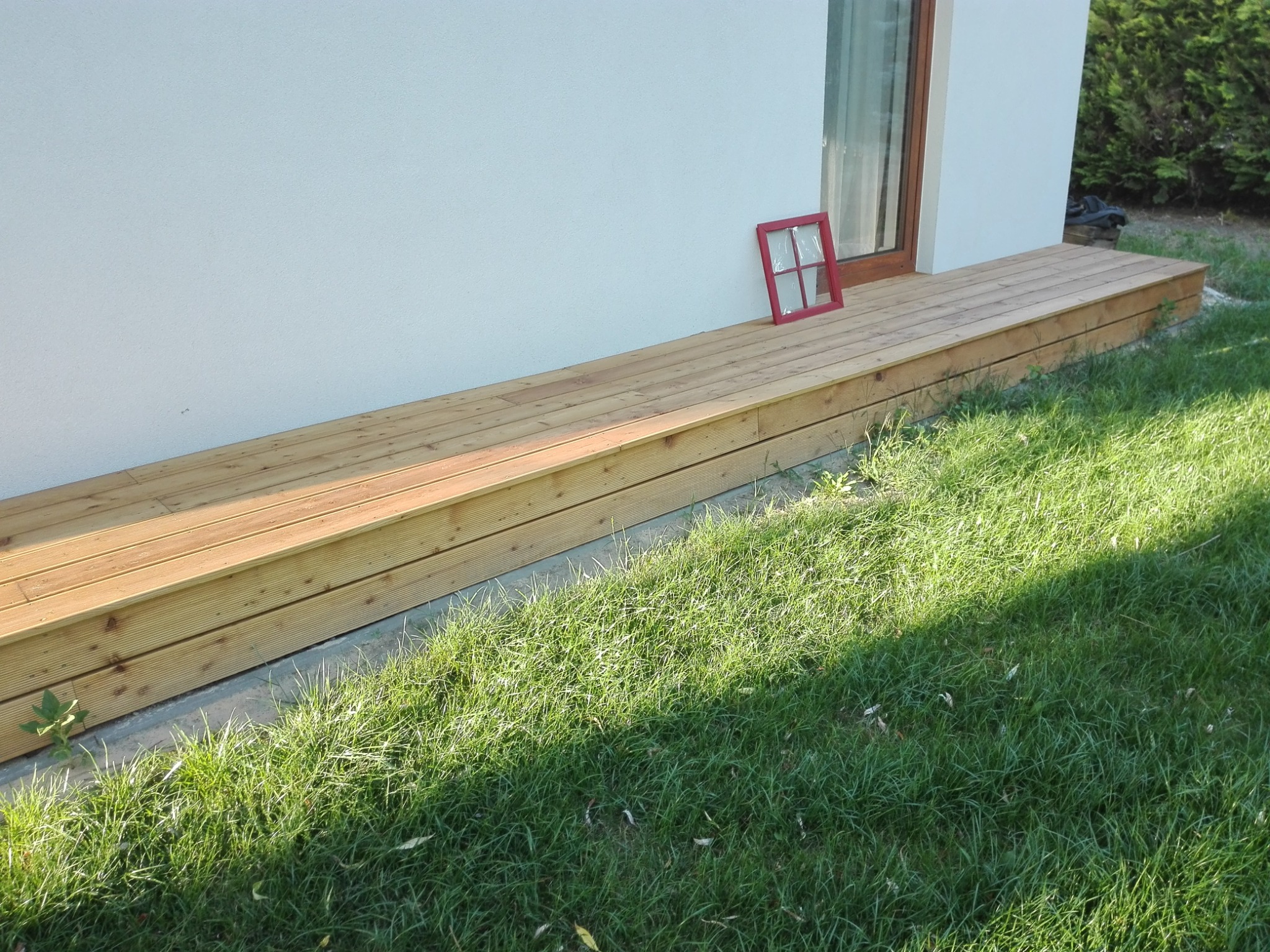 Jasny taras z desek przy białej ścianie domu, z czerwoną ramką okienną opartą o ścianę. Widok na trawnik w słoneczny dzień.
