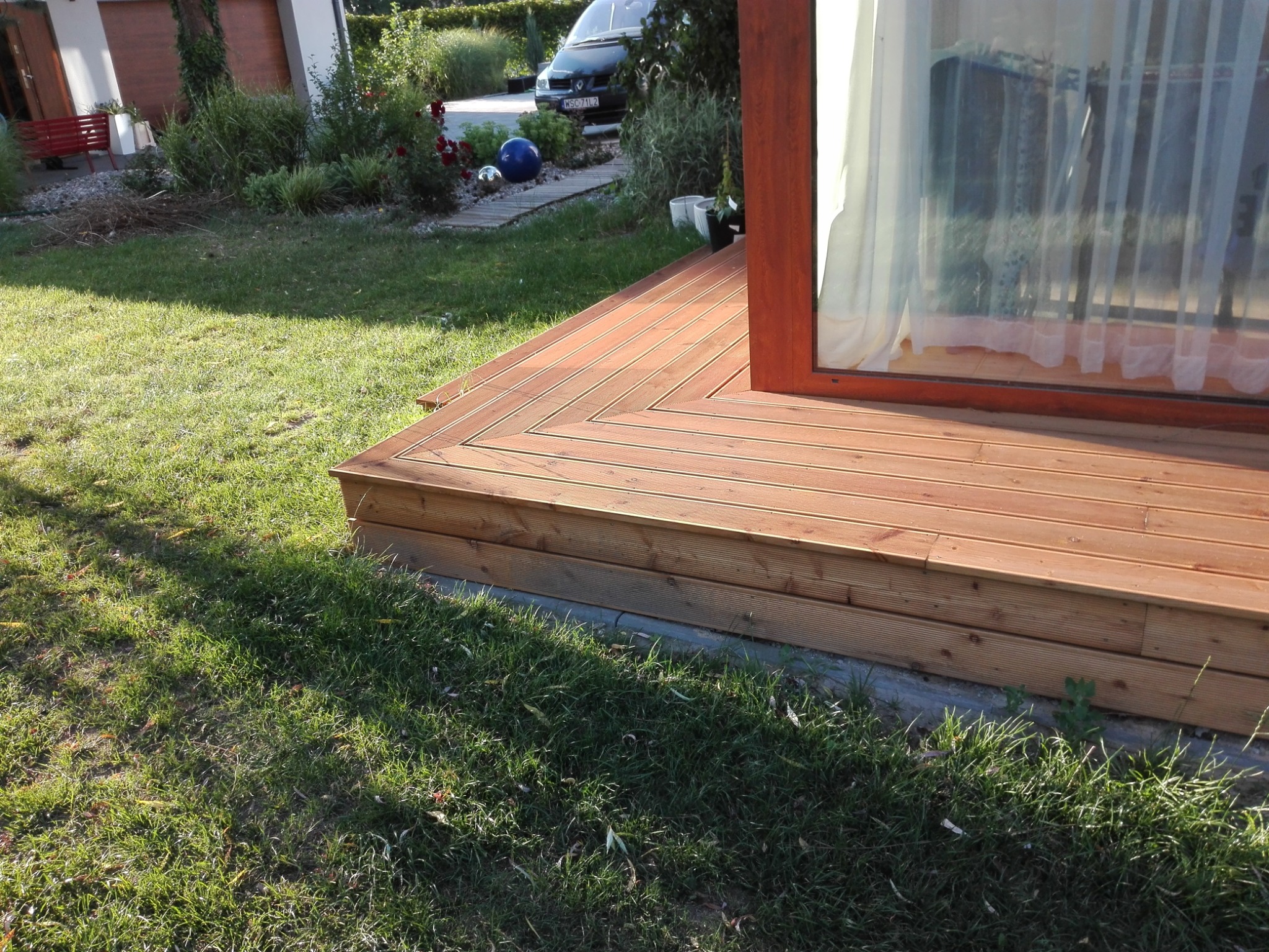 Jasny taras drewniany z desek ułożonych w jodełkę, przylegający do budynku z oknem. W tle trawnik, roślinność i zaparkowany samochód.