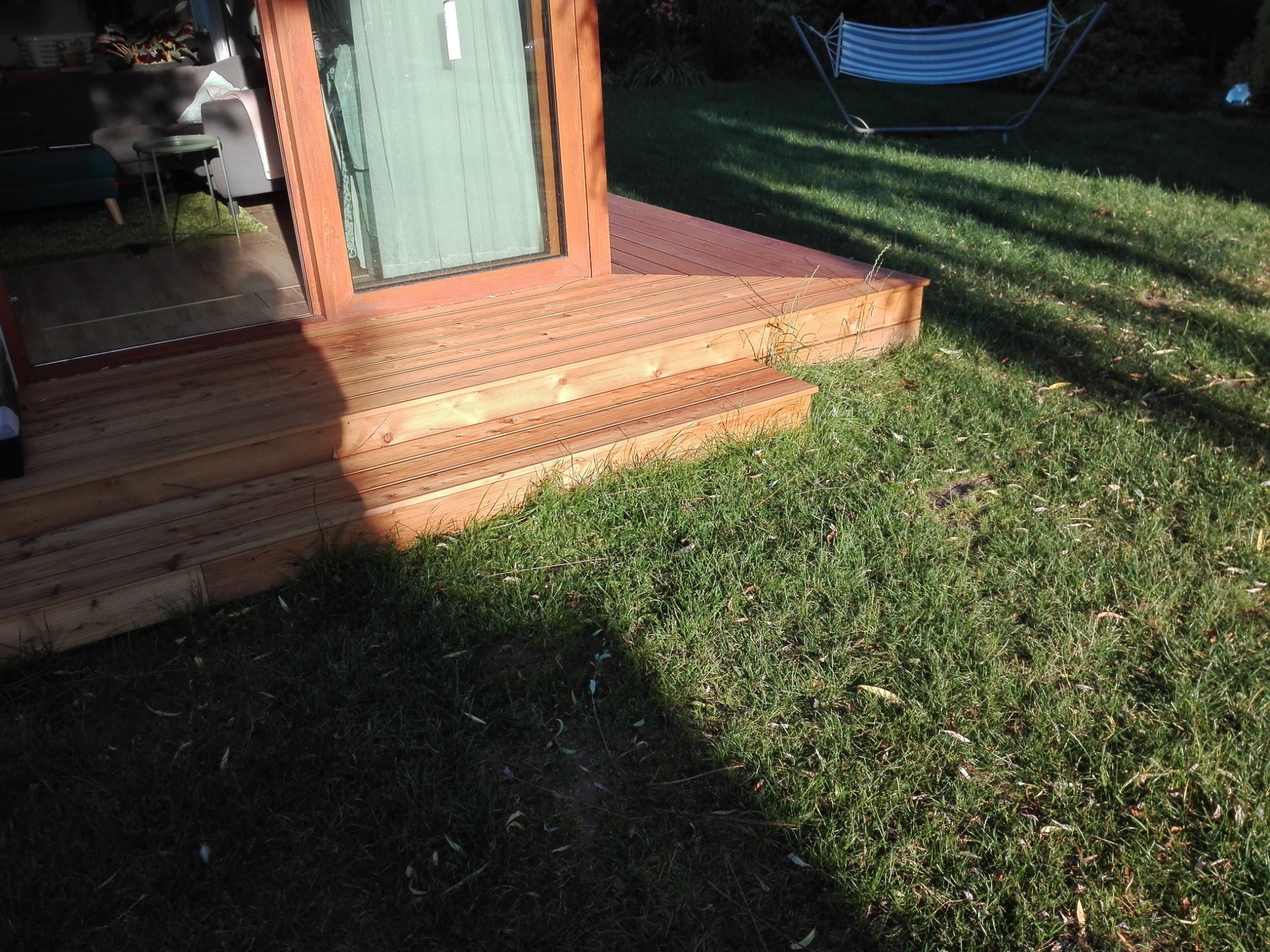 Drewniany taras z niskimi schodami prowadzący do przeszklonych drzwi. W tle trawnik i hamak.