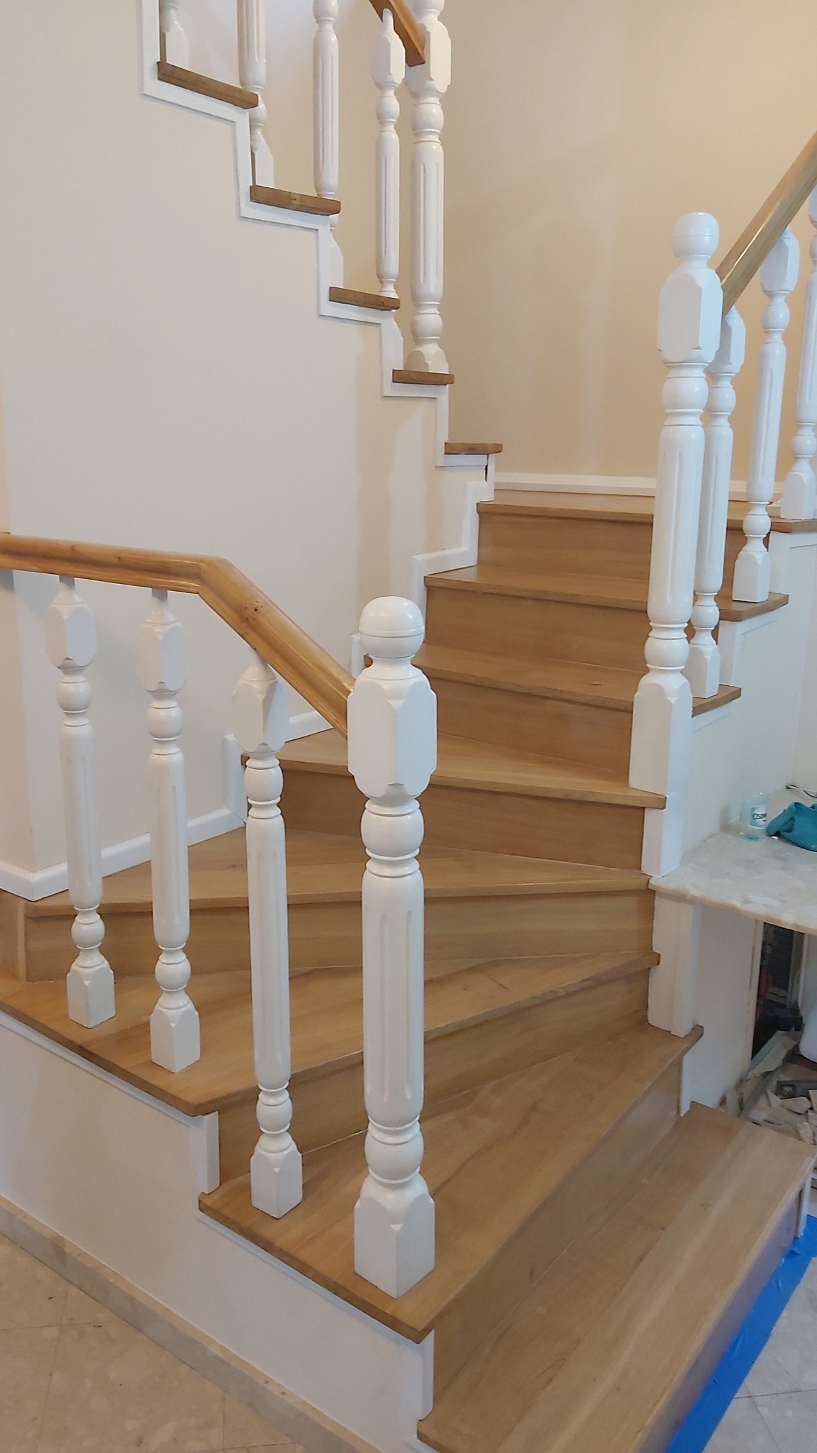 Jasne drewniane schody z białymi balustradami w klasycznym stylu. Widoczne detale wykończenia i naturalne oświetlenie podkreślające strukturę drewna.