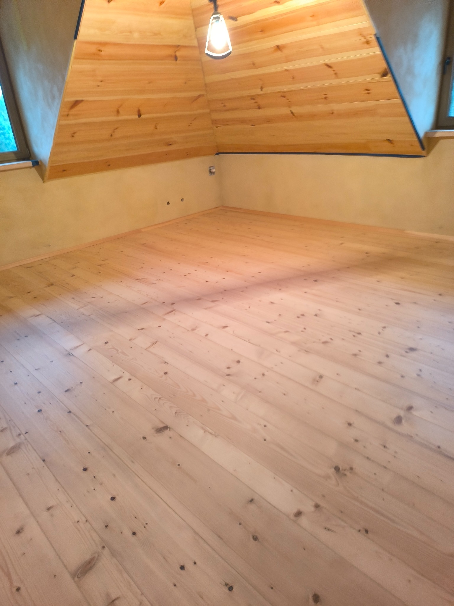 Jasne, drewniane deski podłogowe ułożone w skos w pokoju na poddaszu. Widoczne sęki i naturalny rysunek drewna. Ściany w odcieniach beżu, drewniany sufit ze skosem.