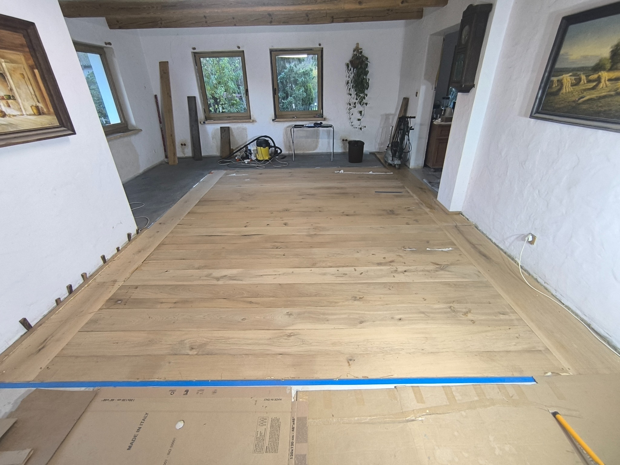 Jasne deski podłogowe w trakcie montażu, zabezpieczone taśmą i kartonem. Widoczne kołki dystansowe przy ścianach. W tle okna i narzędzia.