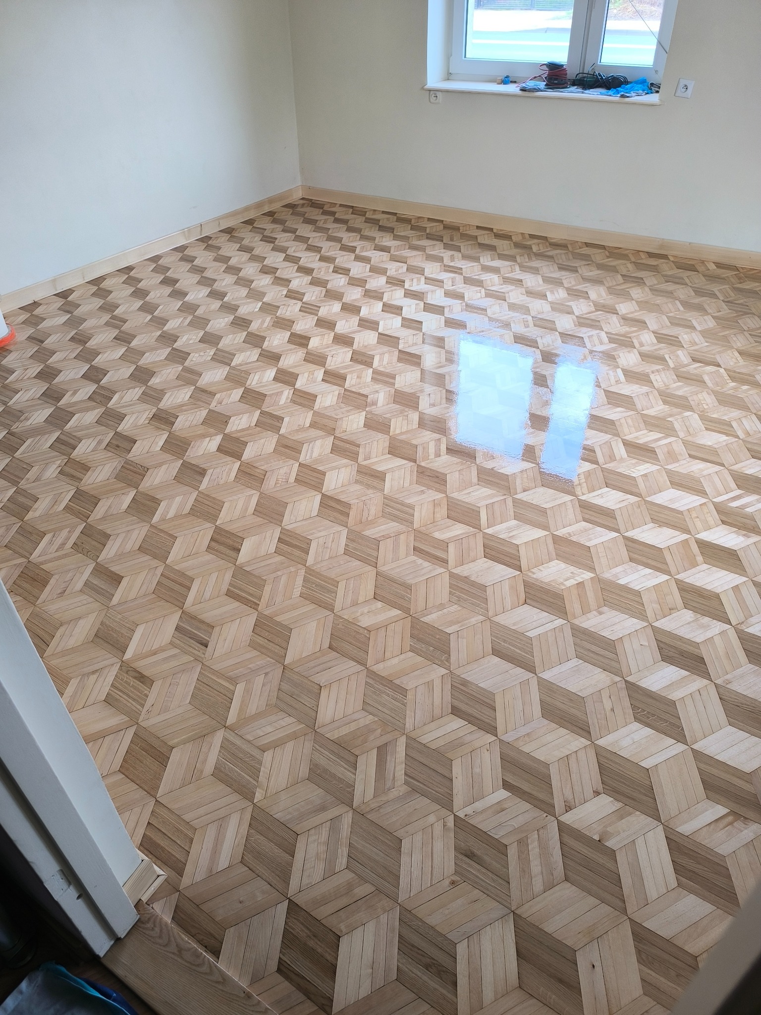 Ukończona podłoga z parkietu ułożonego we wzór 3D w jasnym pokoju z oknem. Widoczne listwy przypodłogowe i odbicie światła na powierzchni podłogi.