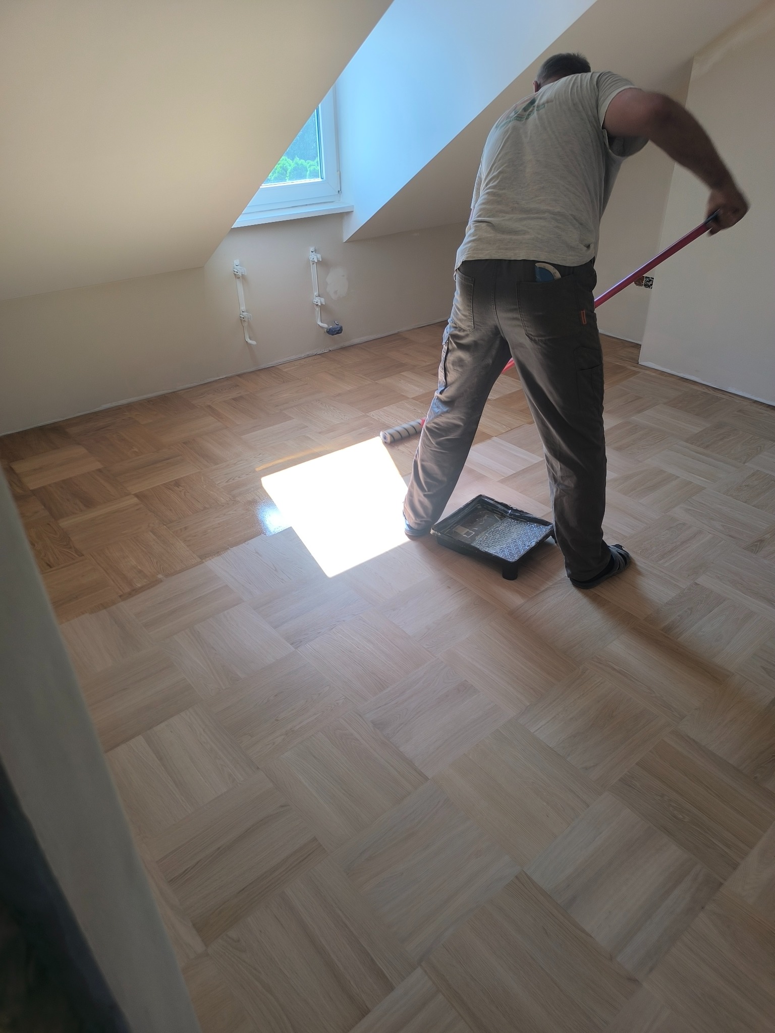 Parkiet w jodełkę po cyklinowaniu, częściowo polakierowany. Widoczny pracownik z wałkiem i kuwetą. Poddasze z oknem dachowym. Jasne, naturalne światło.