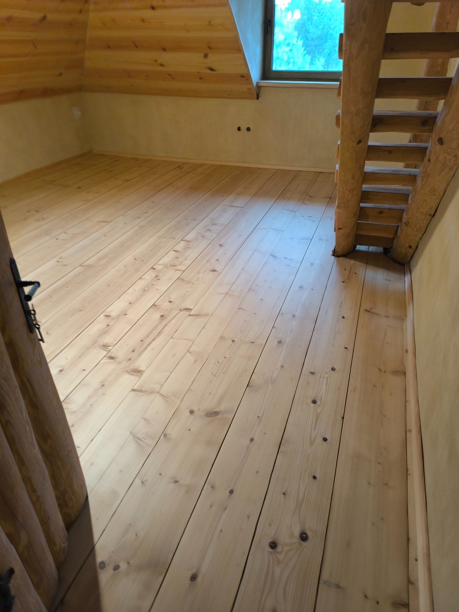 Jasna, drewniana podłoga z widocznymi słojami, ułożona w skos, z fragmentem drewnianych schodów i drzwi po bokach. Naturalne oświetlenie z okna w tle.