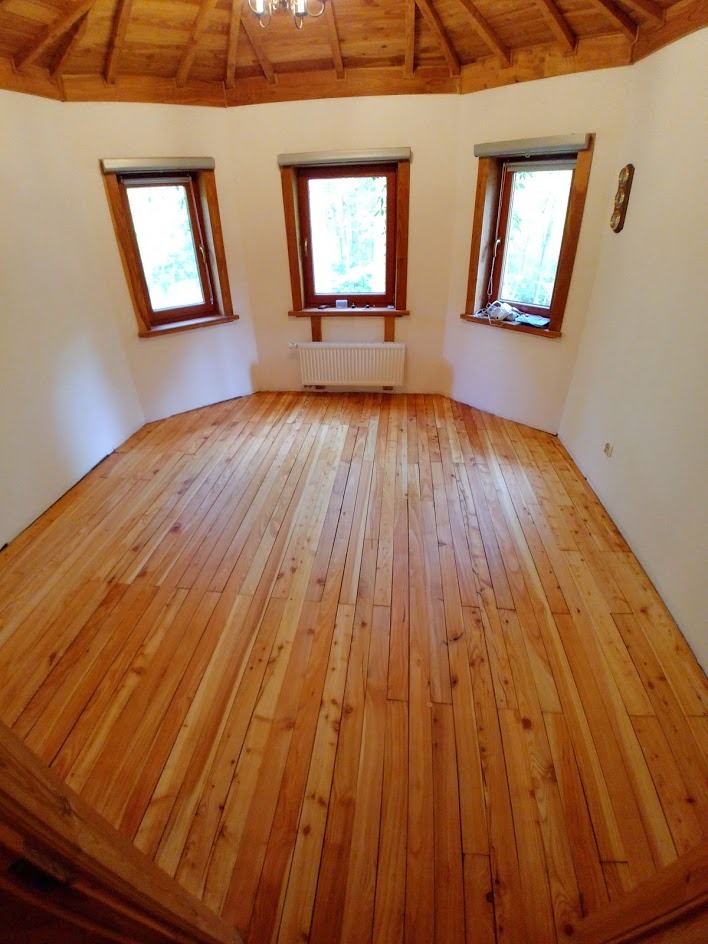 Jasna, drewniana podłoga z desek w okrągłym pokoju z trzema oknami. Widoczny fragment drewnianego sufitu i białe ściany. Naturalne światło.