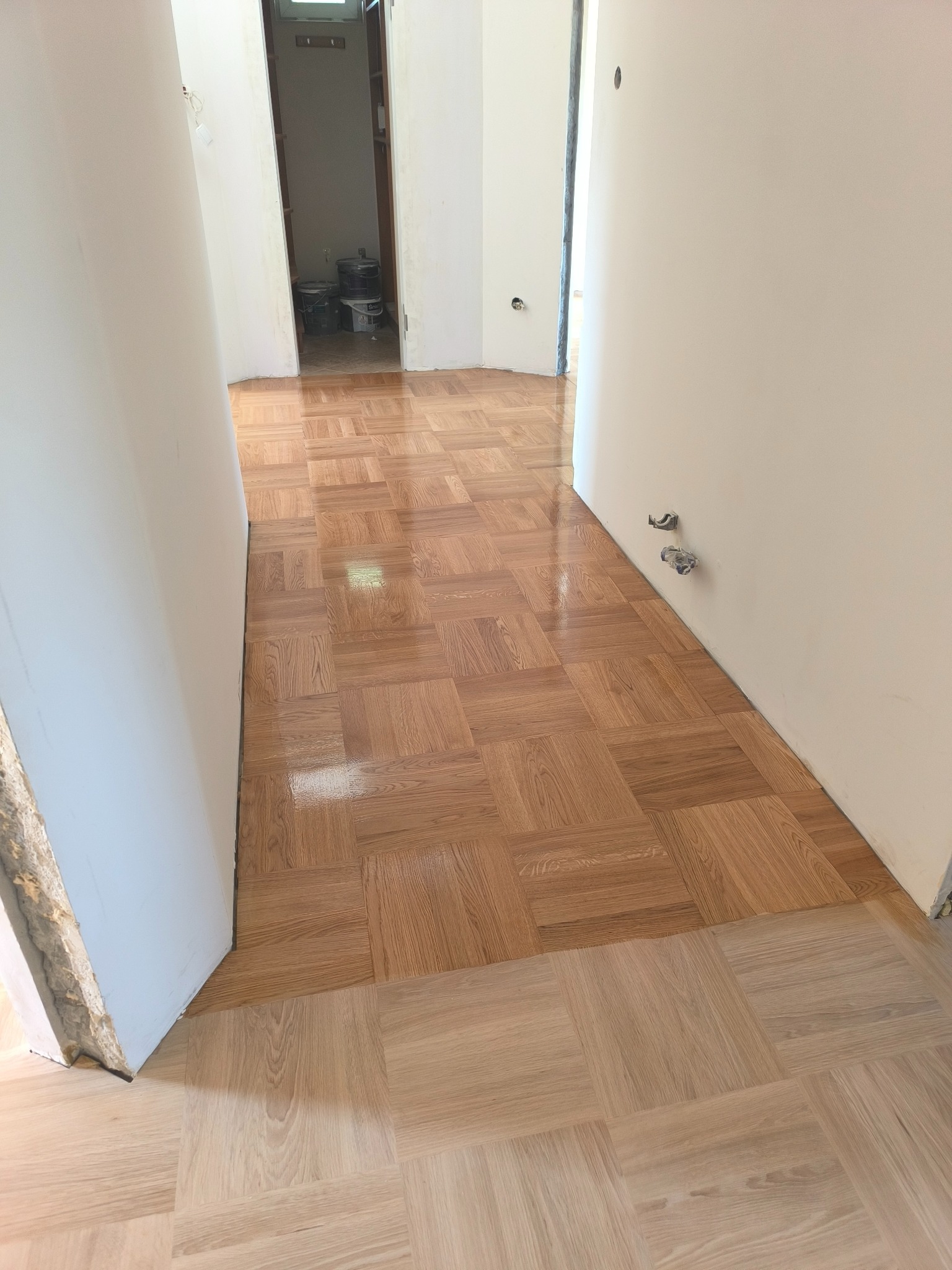 Błyszcząca, nowa podłoga parkietowa ułożona w kwadraty, widoczna w korytarzu z białymi ścianami. Część podłogi jaśniejsza, prawdopodobnie przed cyklinowaniem.