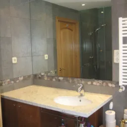 Szara łazienka z marmurowym blatem, umywalką, grzejnikiem drabinkowym i lustrem odbijającym kabinę prysznicową i drewniane drzwi.