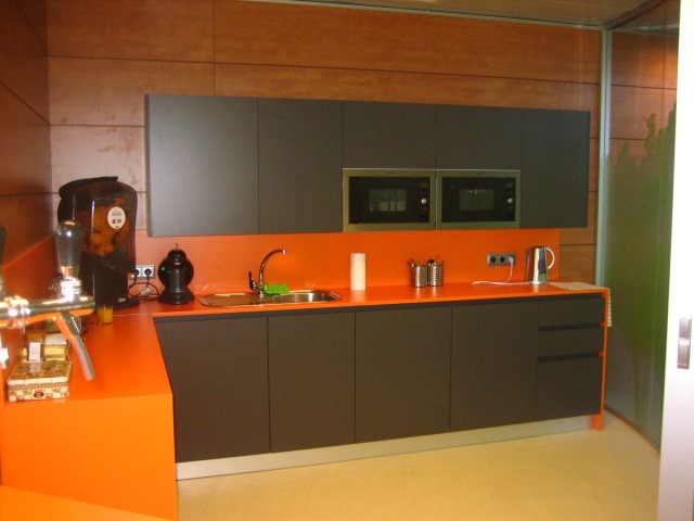 Nowoczesna kuchnia biurowa z pomarańczowym blatem, szarymi szafkami i dwoma wbudowanymi mikrofalówkami, uzupełniona dystrybutorem soku i czajnikiem.