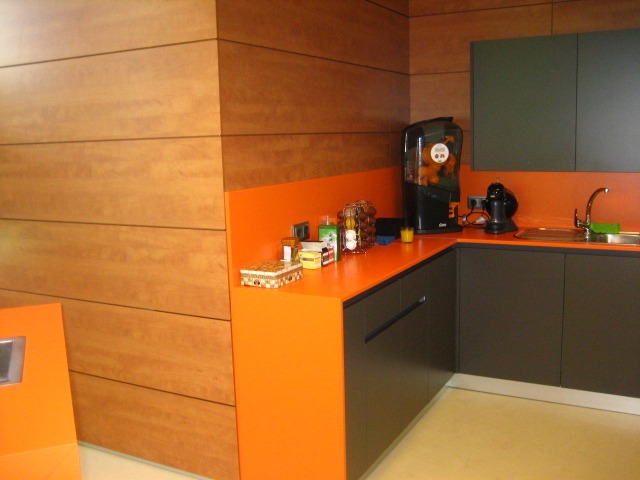 Nowoczesna kuchnia z drewnianą boazerią, pomarańczowym blatem i ciemnoszarymi szafkami, wyposażona w ekspres do kawy i dystrybutor napojów.